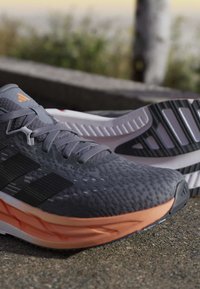 Chaussures de course gris et noir avec dessus texturé et semelles intermédiaires orange posées sur un sol rugueux en extérieur.