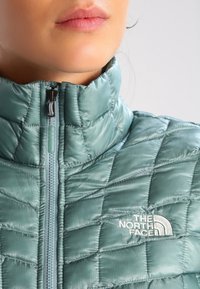 Prošívaná bunda v měkké tyrkysové látce s vysokým límcem, vybavená předním zipem a logem North Face vyšitým na bílo.