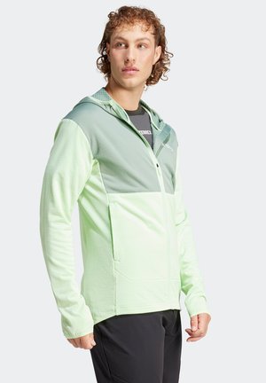  Lichtgroene windbreaker-jack met capuchon, voorzien van een gestructureerde stof, een ritssluiting aan de voorkant en contrasterende donkerdere groene panelen bovenaan.