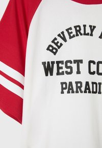 T-shirt blanc à manches courtes avec manches rouges et rayures, avec texte noir "Beverly West Coast Paradise" sur le devant.