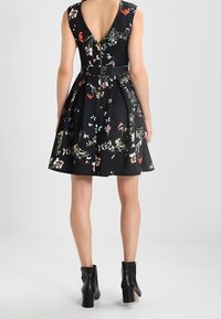 Robe noire à fleurs avec un dos en V, taille cintrée et jupe évasée. Présente des motifs floraux colorés et une fermeture éclair visible. Associée à des bottes à talons noires.