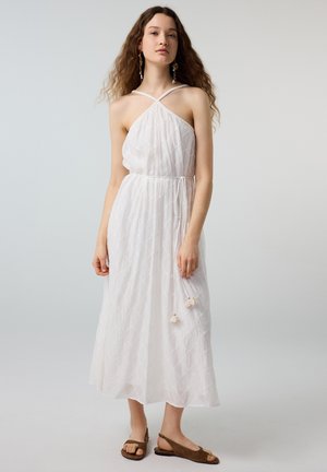 Frau mit langen lockigen Haaren, die ein weißes Halter-Midikleid mit gebundener Taille und braune Sandalen mit offenen Zehen trägt, steht vor einem einfachen Hintergrund.