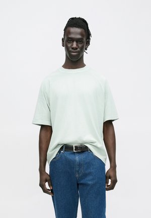 Nuori tummaihoinen mies, jolla on rastat, pukeutuneena väljään vaaleanvihreään T-paitaan, joka on työntynyt sinisiin farkkuihin mustan vyön kanssa, seisoo yksinkertaisen valkoisen taustan edessä.