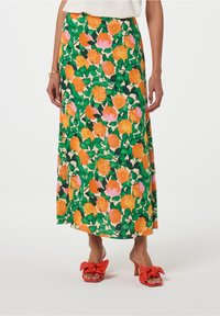 Florale maxi rok met grote oranje en roze rozen op een groene achtergrond, met een vloeiende uitstraling en een split aan de onderrand. Paar rode strik sandalen.