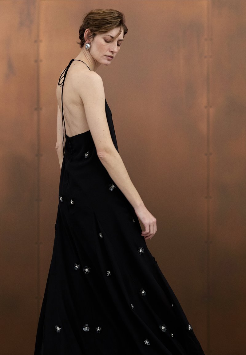 Black sleeveless dress with a low back, featuring silver floral embroidery and a flowing design. Smooth fabric with a slight sheen.