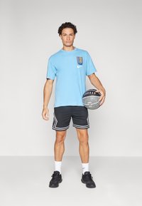 Világoskék pamut póló logóval, fekete sportnadrág fehér csíkkal, és fekete kosárlabda cipő. Egy kosárlabdát tart.