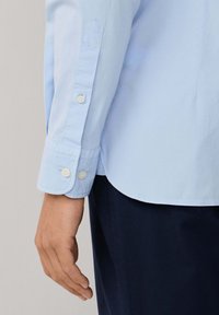 Lichtblauw overhemd met lange mouwen en met knopen gesloten manchetten, met een gladde textuur, een puntige mouwontwerp en een afgeronde zoom. Donkere broek zichtbaar.