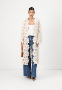 Lace long coat in ivory, featuring floral patterns, open front, and wide sleeves. Paired with a white top and blue wide-leg jeans.