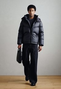 Casaco acolchoado preto com gola alta, fecho de correr e acolchoamento horizontal; combinado com jeans escuros de pernas largas e uma bolsa preta.