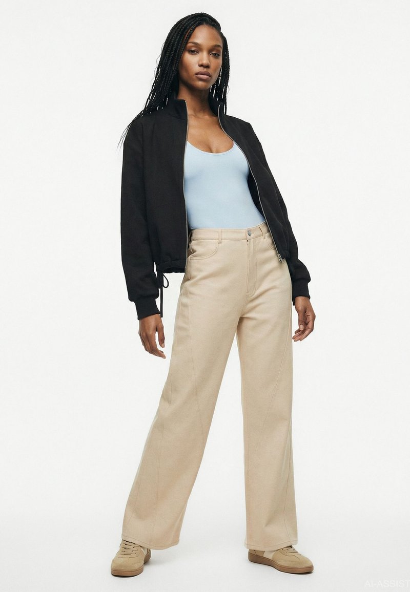 Femme avec de longues tresses portant un débardeur bleu clair, une veste noire zippée, un pantalon beige à jambes larges et des chaussures beiges, debout dans une pose décontractée.