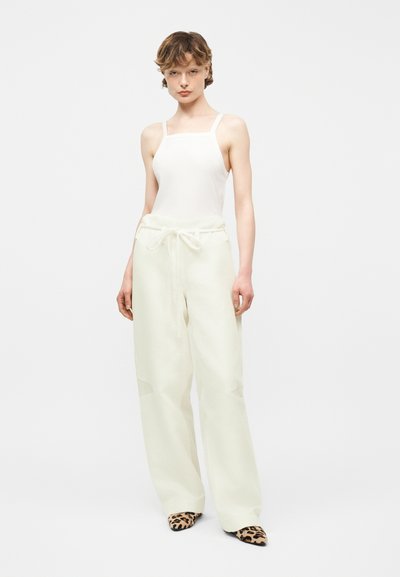 Jeune personne debout portant un haut blanc sans manches, un pantalon blanc ample avec cordon de serrage, et des chaussures à bout pointu imprimé léopard sur un fond blanc.