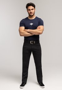 Mężczyzna stojący z założonymi rękami, ubrany w granatowy T-shirt, czarny pasek, ciemnoszare spodnie i czarne trampki na jednolitym tle.