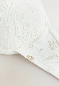 Weiße Spitzen-BH-Schale mit Blumenmustern und einem kleinen goldenen Verschlussdetail auf neutralem Hintergrund.