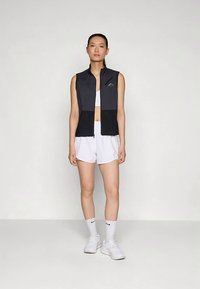 Črni brezrokavnik z zadrgo in teksturiranimi paneli, nošen preko bele kratke majice in hlačk, kombiniran z belimi čevlji in nogavicami.