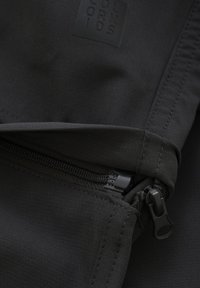 Svart tyg med en slät yta och dragkedjestängning. Detaljer inkluderar en liten präglad logotyp och prydlig sömnad längs kanterna.