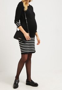 Vestido de maternidad a rayas en blanco y negro con mangas largas, de corte ajustado, combinado con zapatos de charol negros y un bolso cruzado negro.