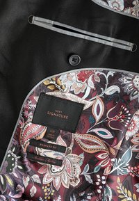Interior de chaqueta de traje negro con patrón cachemir. Presenta una etiqueta que dice "NEXT SIGNATURE" y las instrucciones de cuidado. Tejido texturizado con acentos.