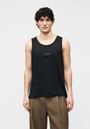 Ung mann med krøllete hår iført en svart ermeløs singlet og løse brune bukser, stående mot en enkel hvit bakgrunn.