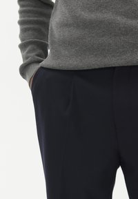 Sivi teksturirani pulover s dugim rukavima, u kombinaciji s tamnoplavim krojenim hlačama s bočnim džepovima i glatkom završnom obradom.