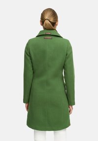 Manteau en mélange de laine vert avec un col, des coutures visibles et deux étiquettes près du col. Le manteau a une silhouette ajustée et un design à longueur de genoux.