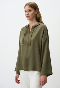 Felpa con cappuccio verde oliva realizzata in tessuto morbido, con una parte anteriore a bottoni, lacci e maniche larghe. Indossata sopra pantaloni bianchi.