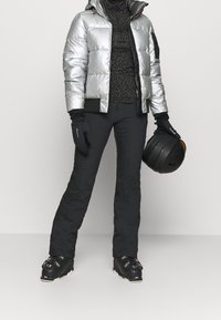 Harmaa hopeanhohtoinen toppatakki hupulla, musta kuvioitu välikerros, mustat housut ja käsineet. Pitelee mustaa laskettelukypärää.