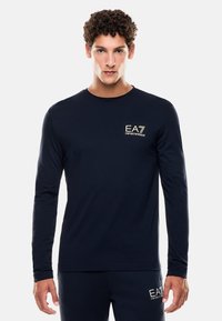 Maglietta a maniche lunghe blu navy realizzata in tessuto morbido, con un piccolo logo dorato EA7 sul petto e un collo rotondo.