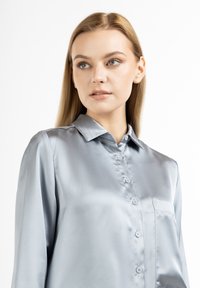 Camicia azzurra in satin con bottoni, maniche lunghe, colletto e taschino sul petto. Texture liscia e leggero lucido.