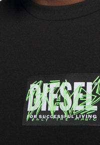 Černé bavlněné tričko s grafickým potiskem v bílé a neonově zelené, na kterém je napsáno "DIESEL" a "PRO ÚSPĚŠNÝ ŽIVOT JEN MAGIE."