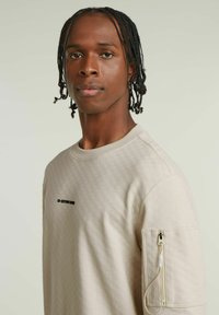 Beżowy sweatshirt z fakturowanym wzorem, okrągłym dekoltem i kieszenią na zamek na rękawie. Posiada czarny logo "G-STAR RAW" na klatce piersiowej.