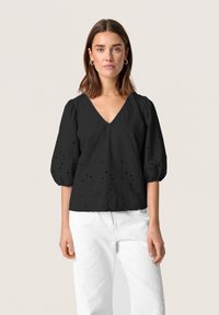 Blusa nera a V con maniche a sbuffo, caratterizzata da un tessuto testurizzato e piccoli buchi ricamati. Abbinata a pantaloni bianchi testurizzati.