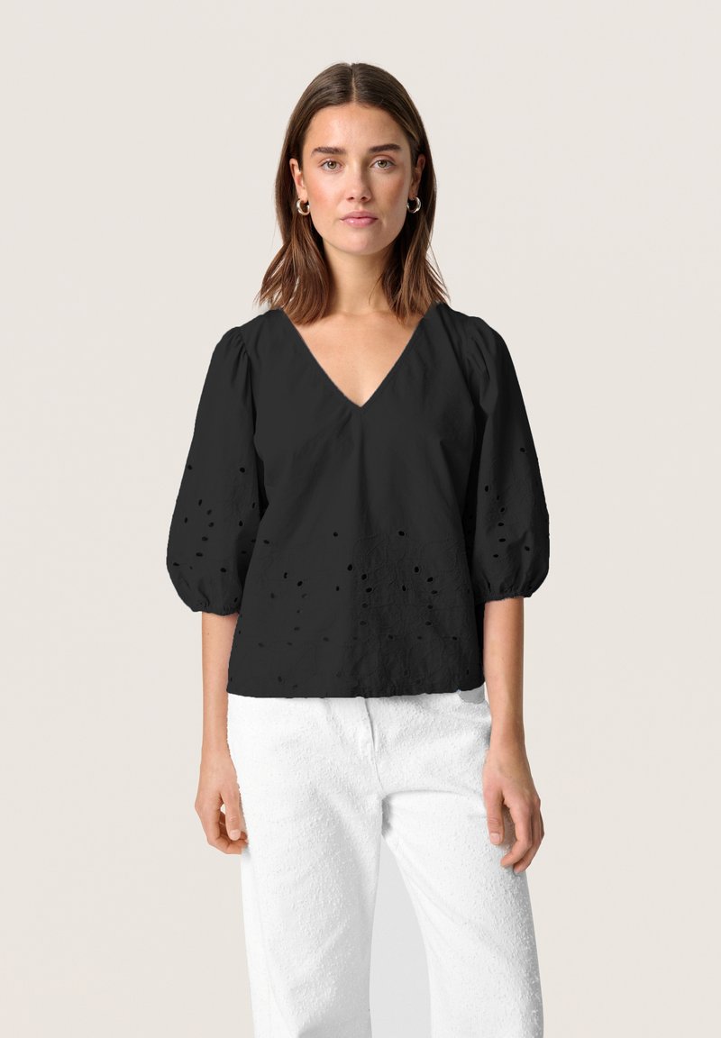 Blusa nera a V con maniche a sbuffo, caratterizzata da un tessuto testurizzato e piccoli buchi ricamati. Abbinata a pantaloni bianchi testurizzati.