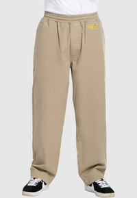 Beige Sweatpants mit elastischem Bund, Vorderfalten und Logodetail. Kombiniert mit schwarzen Sneakern mit weißen Akzenten und Schnürsenkeln.