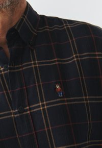 Cămașă în carouri bleumarin cu dungi maro și aurii. Are nasturi închise la culoare și un mic personaj brodat în formă de urs pe piept. Textură moale.