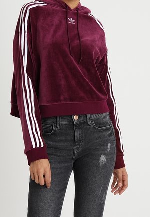 Kobieta w bordowej welurowej bluzie z kapturem Adidas z białymi paskami na rękawach i ciemnych, przetartych dopasowanych jeansach.
