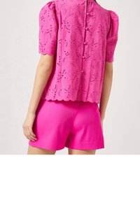 Blusa di pizzo rosa con maniche corte a sbuffo, caratterizzata da un orlo ondulato e chiusura con bottoni, abbinata a pantaloni corti rosa vivace.