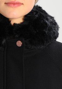 Persona che indossa un cappotto nero con un spesso e morbido colletto di pelliccia nera e un bottone di rame vicino al collo, visibile dalla mascella fino alle spalle.