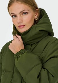 Zelená polstrovaná bunda s vysokým límcem, elastickými manžetami a prošívaným designem. Materiál se zdá být hladký a vodoodpudivý.