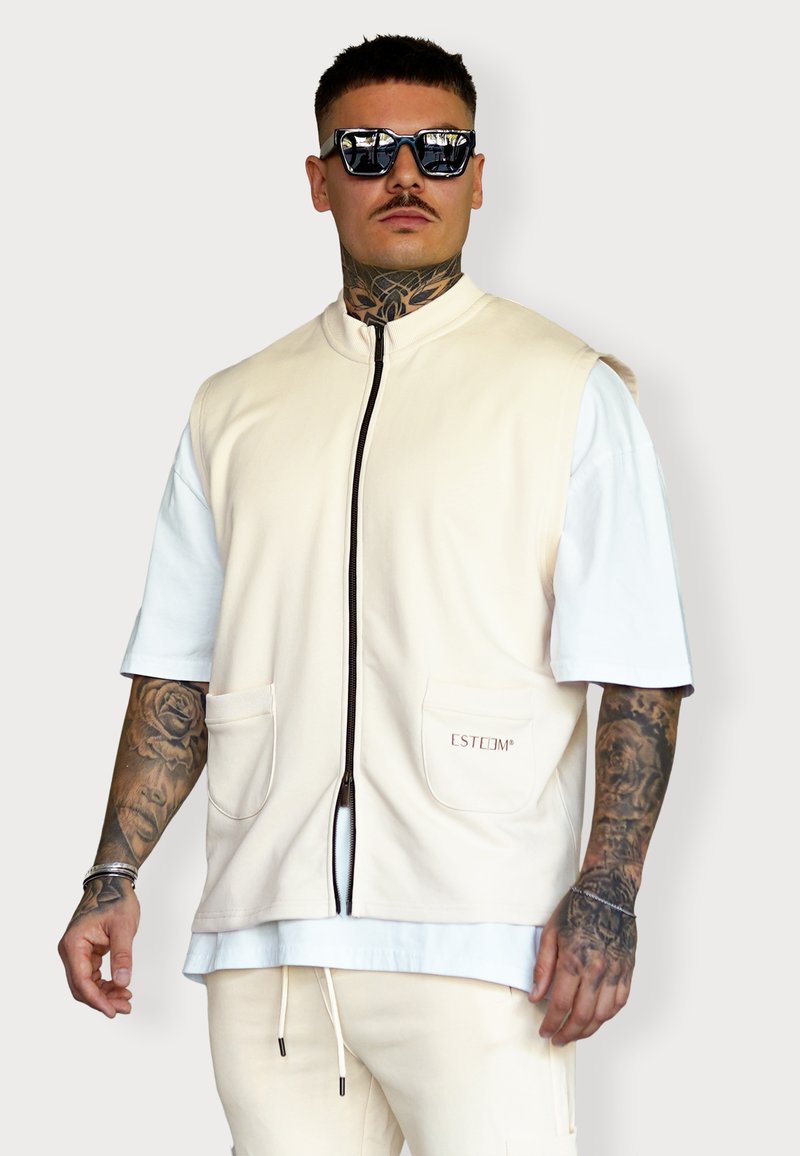 Beige Zip-Up-Weste mit kurzen weißen Ärmeln, zwei Fronttaschen und kleinem Logo-Detail. Das Modell hat Tattoos und trägt reflexionssichere Sonnenbrillen.