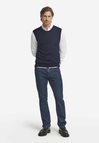 Marinblå stickad väst över en vit skjorta med krage, parat med mörk denimjeans och svarta skor. Enkelt, avslappnat design med minimala accenter.