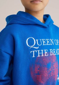 Modra kapuca s napisom "Kraljica ritma" v belem, z rdečo disco kroglo grafiko. Mehak material, kapuco s vrvico in prostoren kroj.