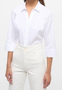 Witte blouse met een kraag, driekwartmouwen en een textuurstof, gecombineerd met hooggepaste lichtgekleurde broeken.