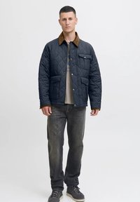 Hombre vistiendo una chaqueta acolchada azul marino con cuello marrón, jeans grises, camiseta gráfica y zapatillas grises, de pie contra un fondo claro.