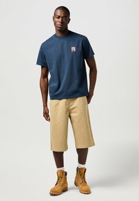 Mörkblå bomulls t-shirt med en liten röd logotyp, i kombination med beige knälånga shorts och bruna snörskor. Enkelt mönster, avslappnad passform.