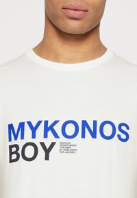 Bílý bavlněný tričko s výrazným modrým a černým textem "MYKONOS BOY" s dalšími menšími nápisy. Kulatý výstřih a standardní střih.