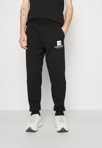 Svarta bomullsoveraller med smalnade ben, försedda med en logotypapplikation och koordinater tryckta på sidan. Mjuk textur och elastiska muddar.