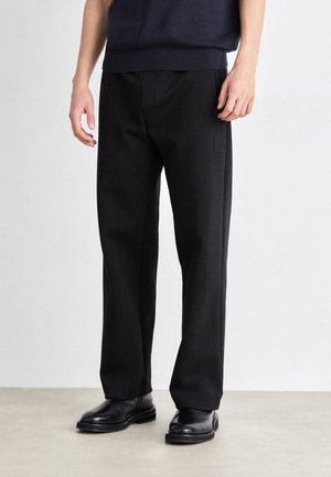 Black tailored trousers with a relaxed fit, featuring a smooth texture and a subtle patch pocket. Worn with black leather boots.
