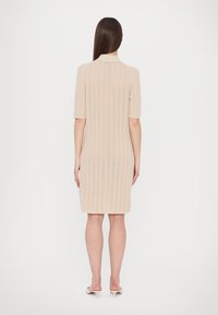 Femme aux longs cheveux bruns porte une robe beige en maille mi-longue et des mules beige à talons, debout de dos sur un fond blanc.