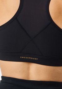 Musta urheilurintaliivit, joissa verkkokangas yläselässä ja kullanvärinen "Swedemount"-logo joustavassa nauhassa, päällä henkilöllä, jolla näkyvissä ihoa selässä.