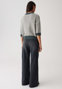 Donna in piedi di spalle, che indossa un maglione a righe verdi e bianche di lana e jeans neri sbiaditi a gamba larga a vita alta.